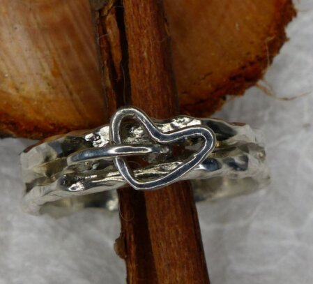 Ring aus 925er Silber mit lebhafter, wilder Oberflaeche, mit eingearbeitetem Spalt, der zur Haelfte des Ringumfangs offen ist. In dem Durchbruch ist eine Oese installiert, in der ein frei schwingendes Herz haengt. Ringbreite 4 - 5 mm. Ringgroeße 58 und 59. Einzelstueck.